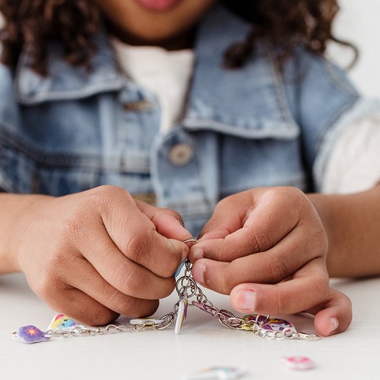 Close-up van kinderhanden die een bedel aan een zilveren ketting bevestigen, met losse bedels.
