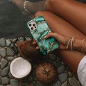 Vrouw houdt een telefoonhoes met groen en goud marmerpatroon vast, naast kokosnoten bij water.