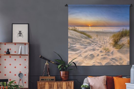 Wandkleed met strandgezicht bij zonsondergang, hangend aan een zwarte roede boven een dressoir in een donkere kamer.