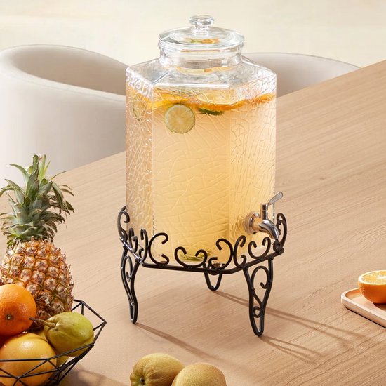 Transparante glazen drankdispenser gevuld met water en fruit, op een zwarte metalen standaard, naast een fruitmand op een houten tafel.