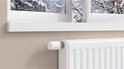 Witte radiatorthermostaat op een witte radiator, met een winterse scène door het raam.