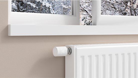 Witte radiatorthermostaat op een witte radiator, met een winterse scène door het raam.