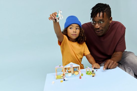 Enfant avec bonnet bleu tenant un cheval LEGO en l'air, père regardant la clinique vétérinaire LEGO.