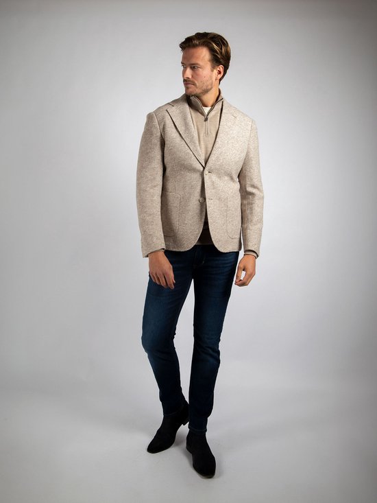 Man in beige colbert met kleine ruitjes, beige trui met rits en donkerblauwe jeans.