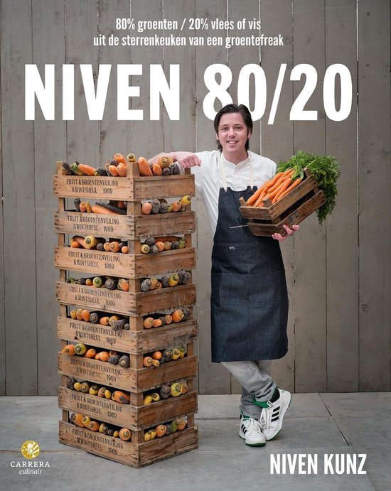 Een man in een koksbuis staat naast een stapel houten kisten vol wortels en andere groenten. Hij houdt een tros wortels vast.