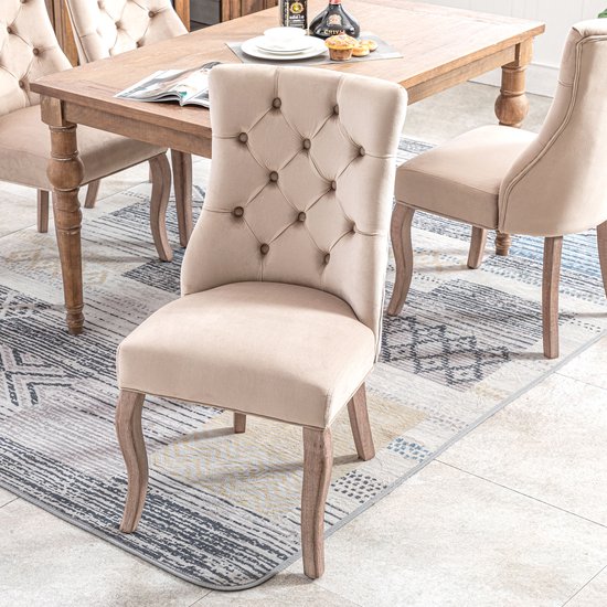 Beige fluwelen eetkamerstoel met knopen-quilting en gebogen houten poten op een vloerkleed.