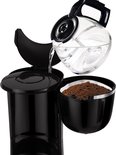 Une main verse de l'eau d'une verseuse en verre dans une cafetière noire contenant du marc de café.