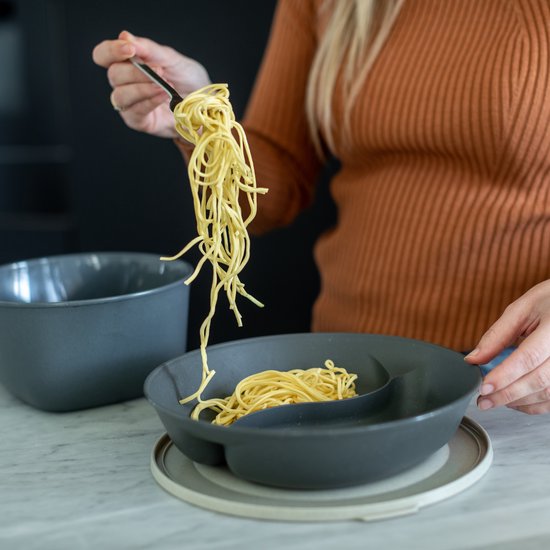 Vrouw met oranje trui schept spaghetti uit een donkergrijs, verdeeld bord.