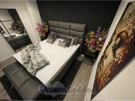 Grijze boxspring met gestoffeerd hoofdbord, luxe kussens, grijze deken en opbergbank.