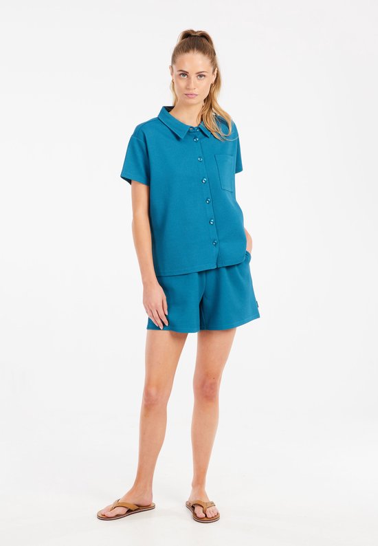 Model met een turquoise korte broek en bijpassend shirt met korte mouwen, staand.