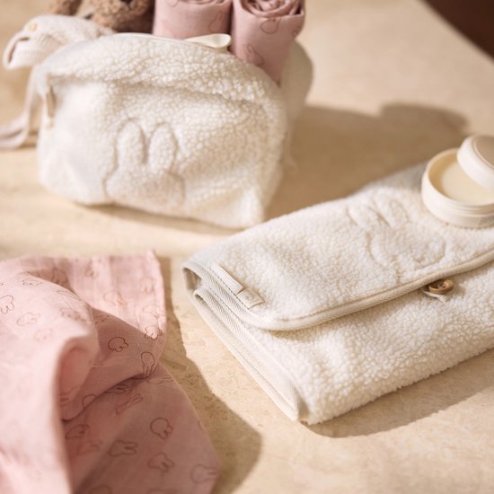 Close-up van een crèmekleurige teddy verschoonmat met beige rand en een houten knoopje. Naast een roze doekje met Nijntje print.