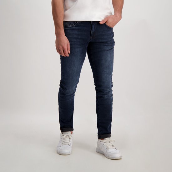 Donkerblauwe, slim fit jeans met lichte slijtage. Een man draagt de jeans met witte sneakers.