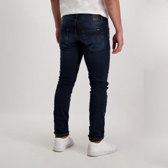 Donkerblauwe, slim fit jeans met een licht verweerde look. Gedragen door een man met witte sneakers.