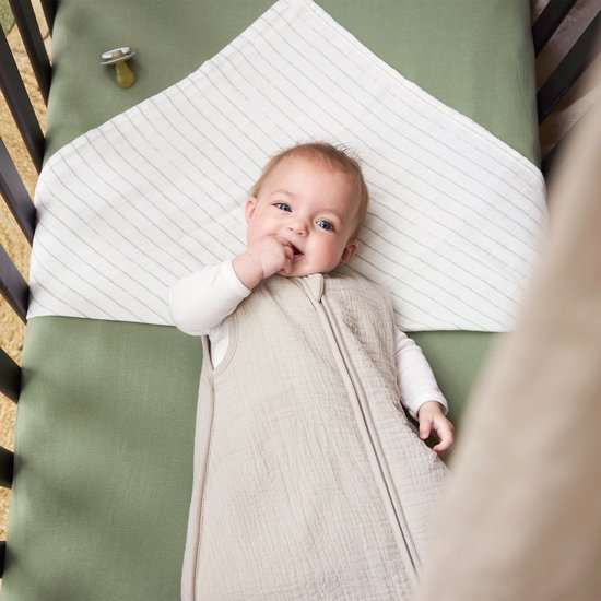 Baby ligt in een wieg in een beige slaapzak, met duim in mond.