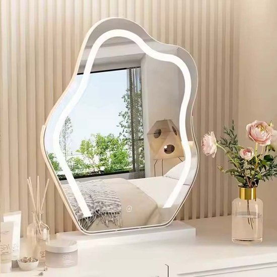 Golvende spiegel met ledverlichting, geplaatst op een kaptafel met cosmetica en een vaas met bloemen.