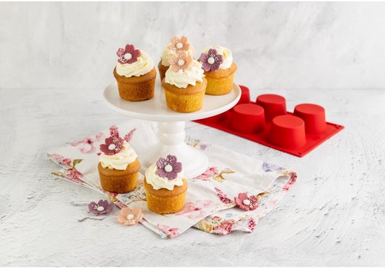 Zes cupcakes met witte crème en bloemen op een witte taartschaal en op een bloemetjesdoek, naast rode muffinvormen.
