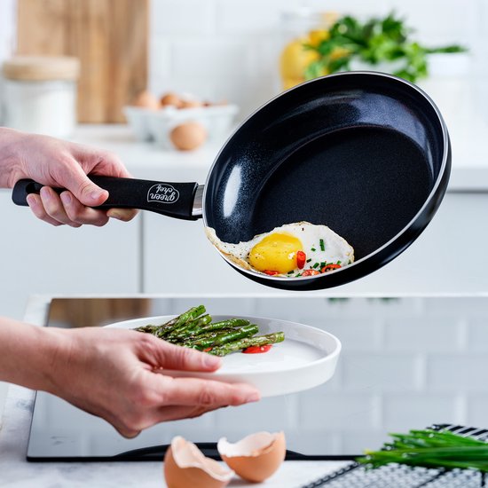 Een gebakken eitje met bosui wordt uit een zwarte koekenpan op een wit bord geschoven. Gebakken asperges liggen op het bord.