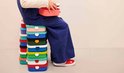 Un enfant en salopette est assis sur une pile de boîtes à lunch Mepal colorées, tenant une boîte à lunch rose.