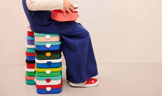 Un enfant en salopette est assis sur une pile de boîtes à lunch Mepal colorées, tenant une boîte à lunch rose.