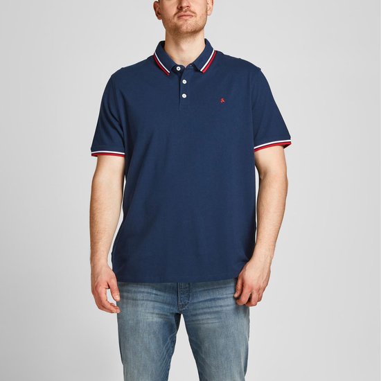 Donkerblauwe polo met rood-wit gestreepte kraag en mouwboorden. Rood logo op borst. Man staat met handen in broekzakken.