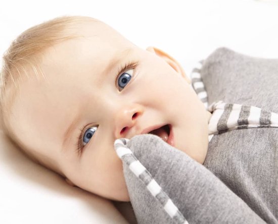 Close-up van een baby met blauwe ogen, die op een witte ondergrond ligt en met de vingers in de grijze Swaddle UP slaapzak bijt.