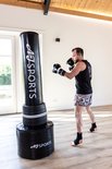 Man in zwarte tanktop en korte broek met bokshandschoenen staat bij zwarte AJ-Sports bokspaal.