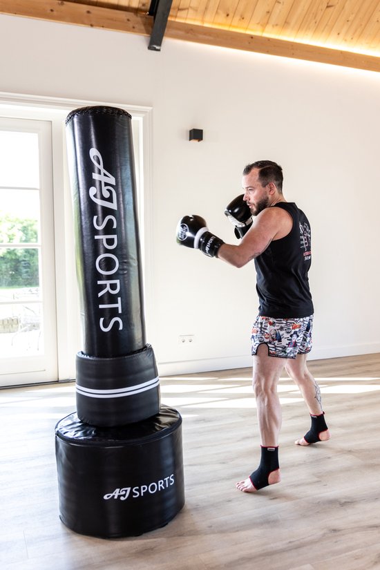 Man in zwarte tanktop en korte broek met bokshandschoenen staat bij zwarte AJ-Sports bokspaal.