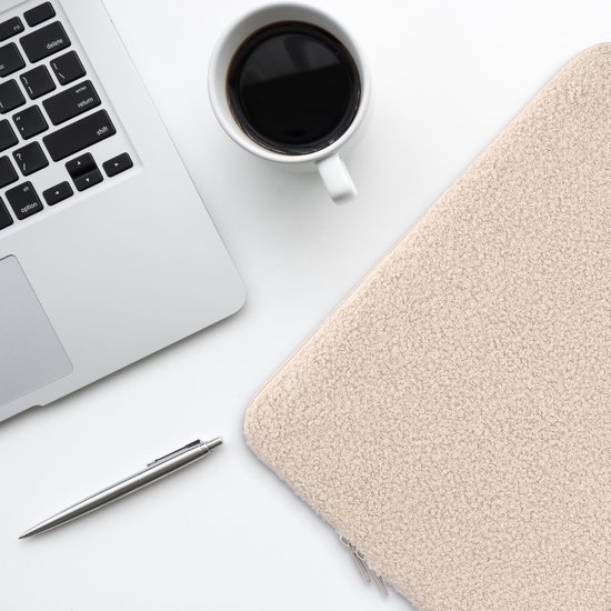 Une housse d'ordinateur portable en peluche beige est posée à côté d'un ordinateur portable, d'une tasse à café et d'un stylo sur un bureau blanc.