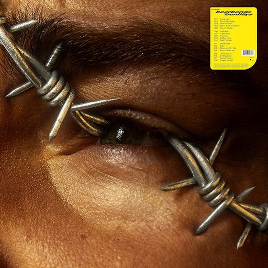 Close-up van een bruin oog bedekt met prikkeldraad, met gele hoes met tracklist.