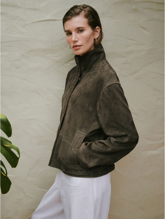 Model draagt een donkergroen suède jack met trechterhals en witte broek.