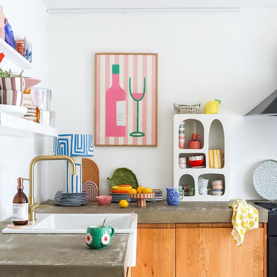 Keuken met witte kasten, aanrechtblad van beton, roze en witte gestreepte poster van een fles en glas, en gekleurd servies.