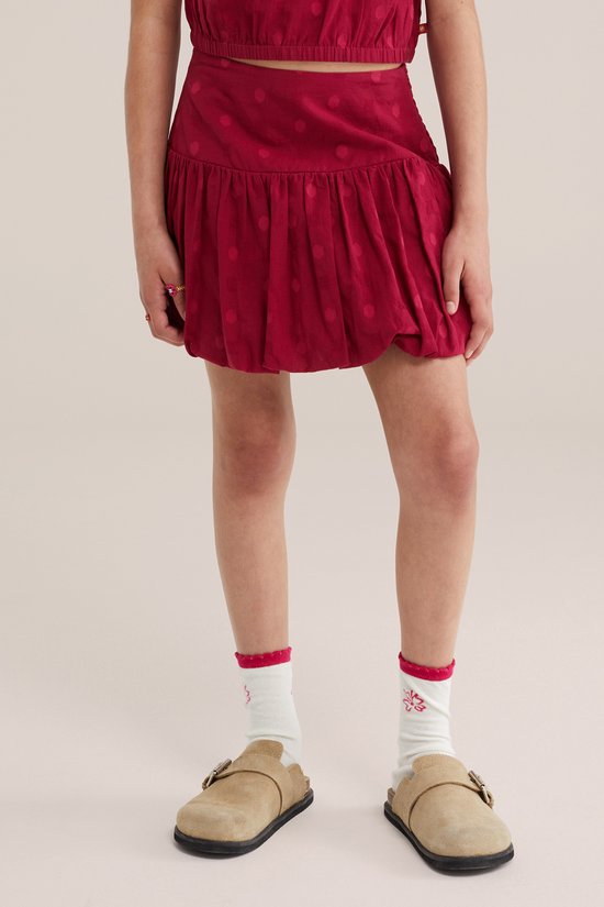 Een meisje draagt een donkerrode minirok met stippen en ruches, met witte sokken en sandalen.