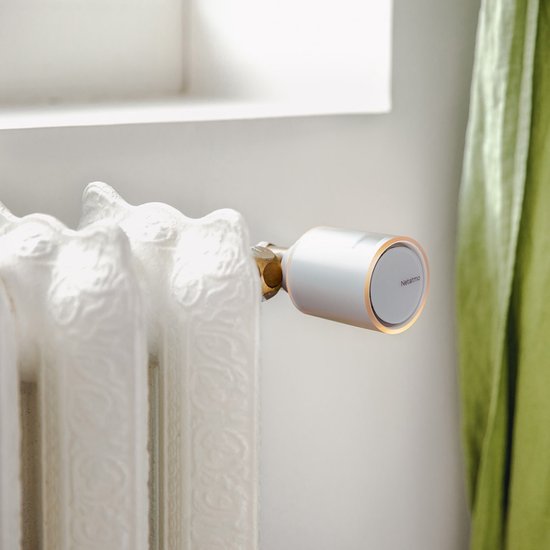 Netatmo slimme thermostaatknop, zilverkleurig met oranje accent, op een crèmekleurige gietijzeren radiator.