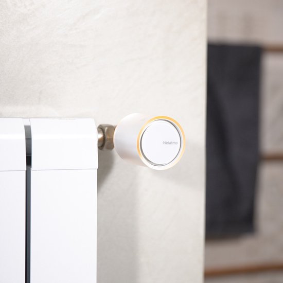 Netatmo slimme thermostaatknop, wit met oranje accent, op witte radiator.