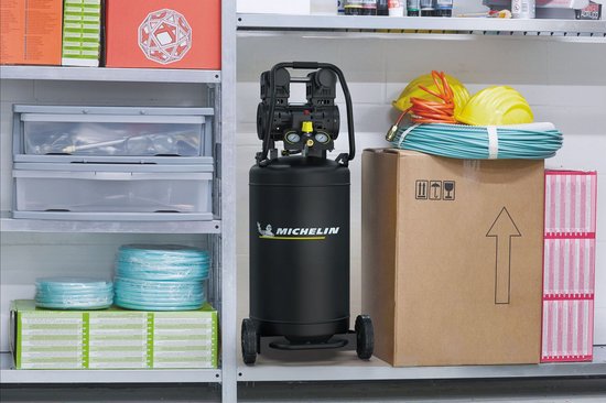 Zwarte staande Michelin compressor met twee wielen, in een werkplaats op een plank naast dozen en gereedschap.