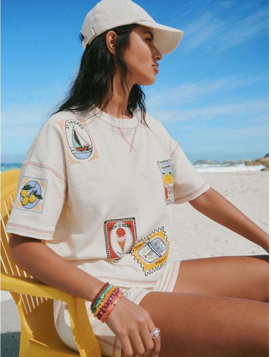 Vrouw in crème T-shirt met vakantie-emblemen en een bijpassende pet op het strand.