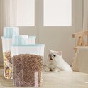 Transparante voorraadbak gevuld met hondenbrokken, met een witte kat naast de bak.