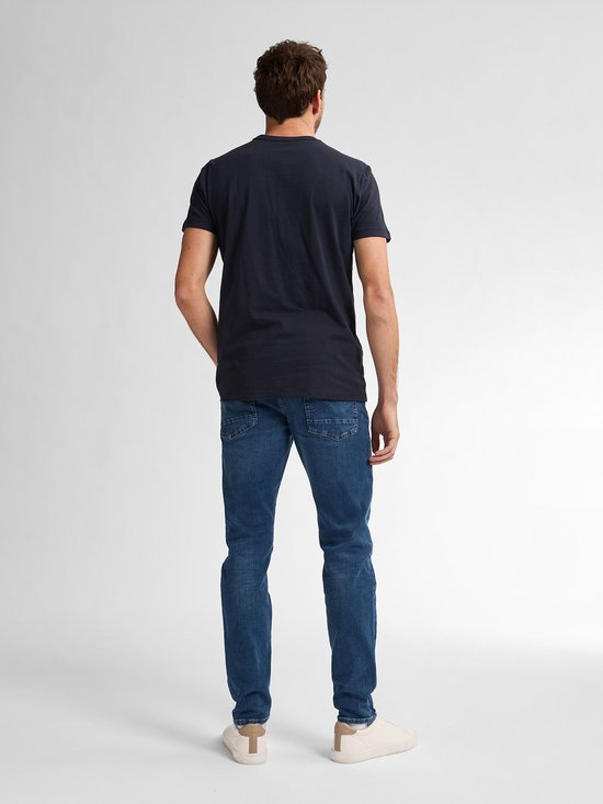 Man in donkerblauw T-shirt en blauwe spijkerbroek, staand met hand in zak.