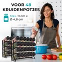 Vrouw met schort die specerijen uit een potje strooit in een keuken, met een rek van 48 potjes op de voorgrond.