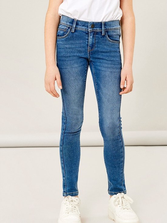 Een meisje draagt een blauwe skinny jeans met witte sneakers, de voorkant van de jeans is zichtbaar.