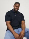 Man in donkerblauw poloshirt met navy kraag, witte knopen en geborduurd "&" logo, zittend op een krukje.