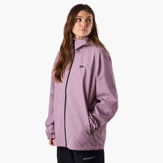 Een vrouw met lang bruin haar in een lichtpaarse windjack met zwarte rits en zwarte broek, handen in de zakken.