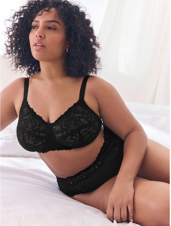Model met krullend haar draagt een zwarte kanten beha en slip, liggend op een wit bed.