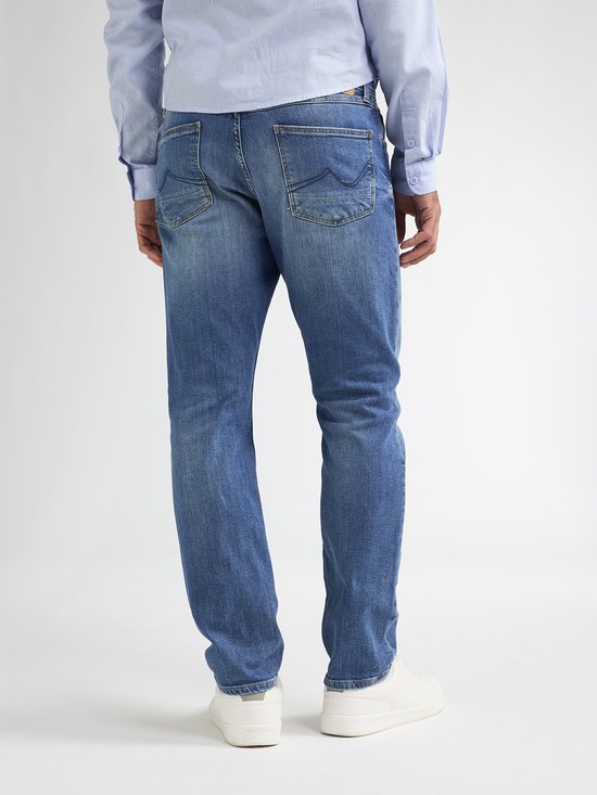 De achterkant van een man in een lichtblauwe spijkerbroek en witte sneakers. Het model draagt een lichtblauwe blouse.