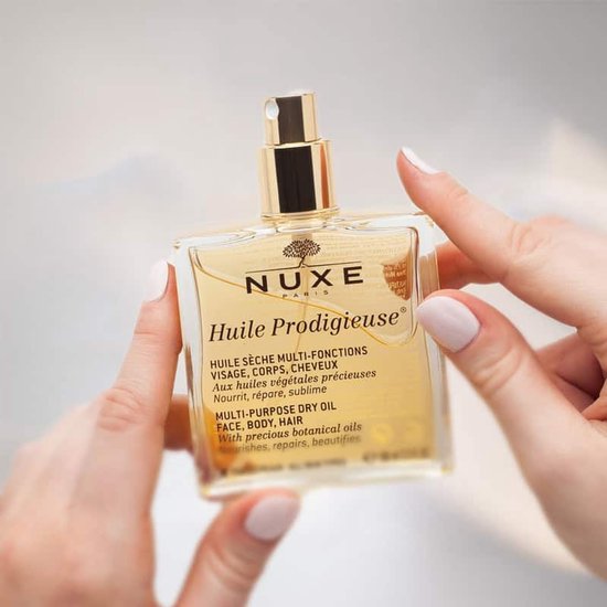 Gouden Nuxe Huile Prodigieuse multi-purpose dry oil in een glazen fles, vastgehouden door twee handen.