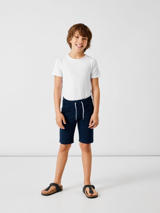 Jongen in een witte T-shirt en donkerblauwe shorts met trekkoord, opgerolde broekspijpen en sandalen.