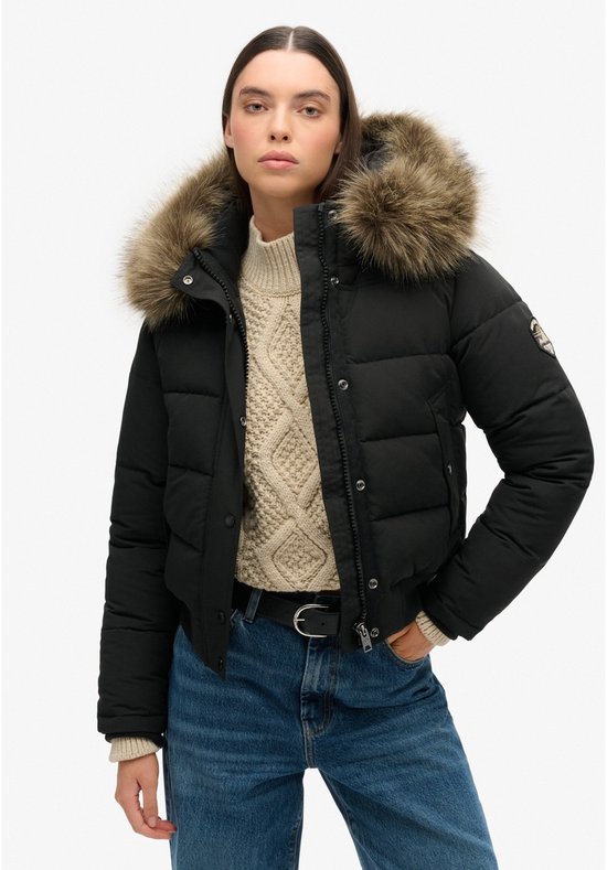 Model in een zwarte korte gewatteerde jas met bontkraag, crème kabeltrui en spijkerbroek.