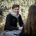 Man met grijze gebreide colsjaal, zittend aan tafel met mok.