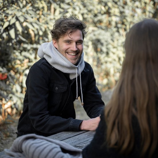 Man met grijze gebreide colsjaal, zittend aan tafel met mok.