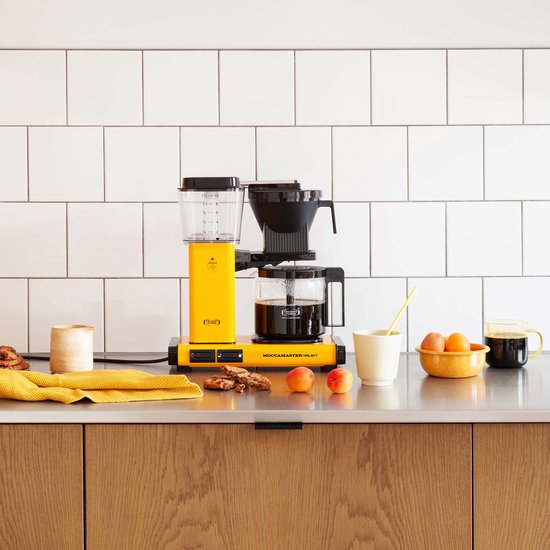 Felgeel Moccamaster koffiezetapparaat met glazen kan gevuld met koffie, naast fruit en koekjes op een aanrecht.